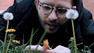 Film still from 'Motion them with breath' (2016) by Jon Beaton. Close-up of a man in his 20s lying in the grass on a grey day. He is surrounded by dandelions and dandelion clocks. In his hand is a dandelion.